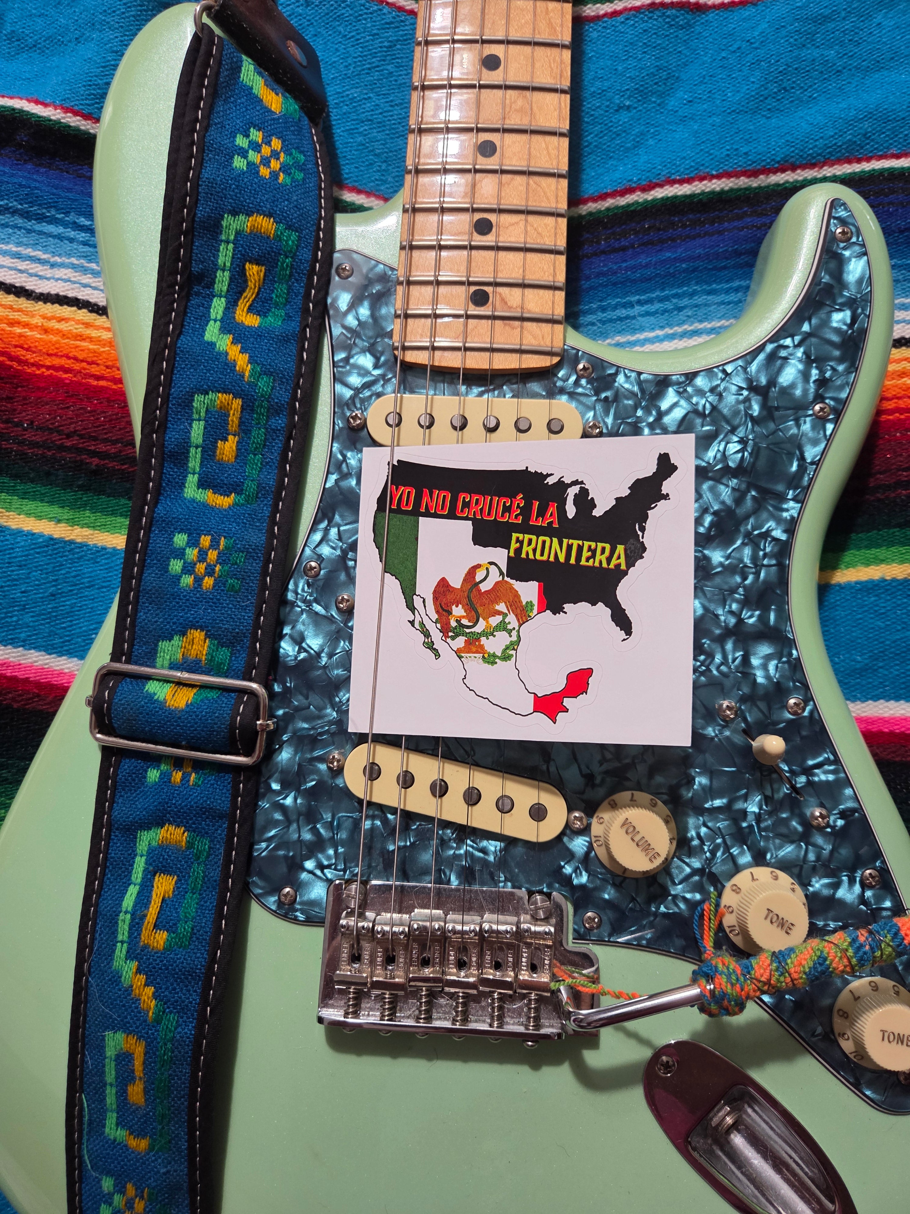 "4" × 4" sticker – Yo No Crucé La Frontera vintage Mexican flag design sticker from 1848 with a close-up of a light green electric Fender Stratocaster guitar with a blue marbled pickguard, a patterned Mexican blue guitar strap, and a small printed sticker that reads 'YO NO CRUCE LA FRONTERA' placed on the body."