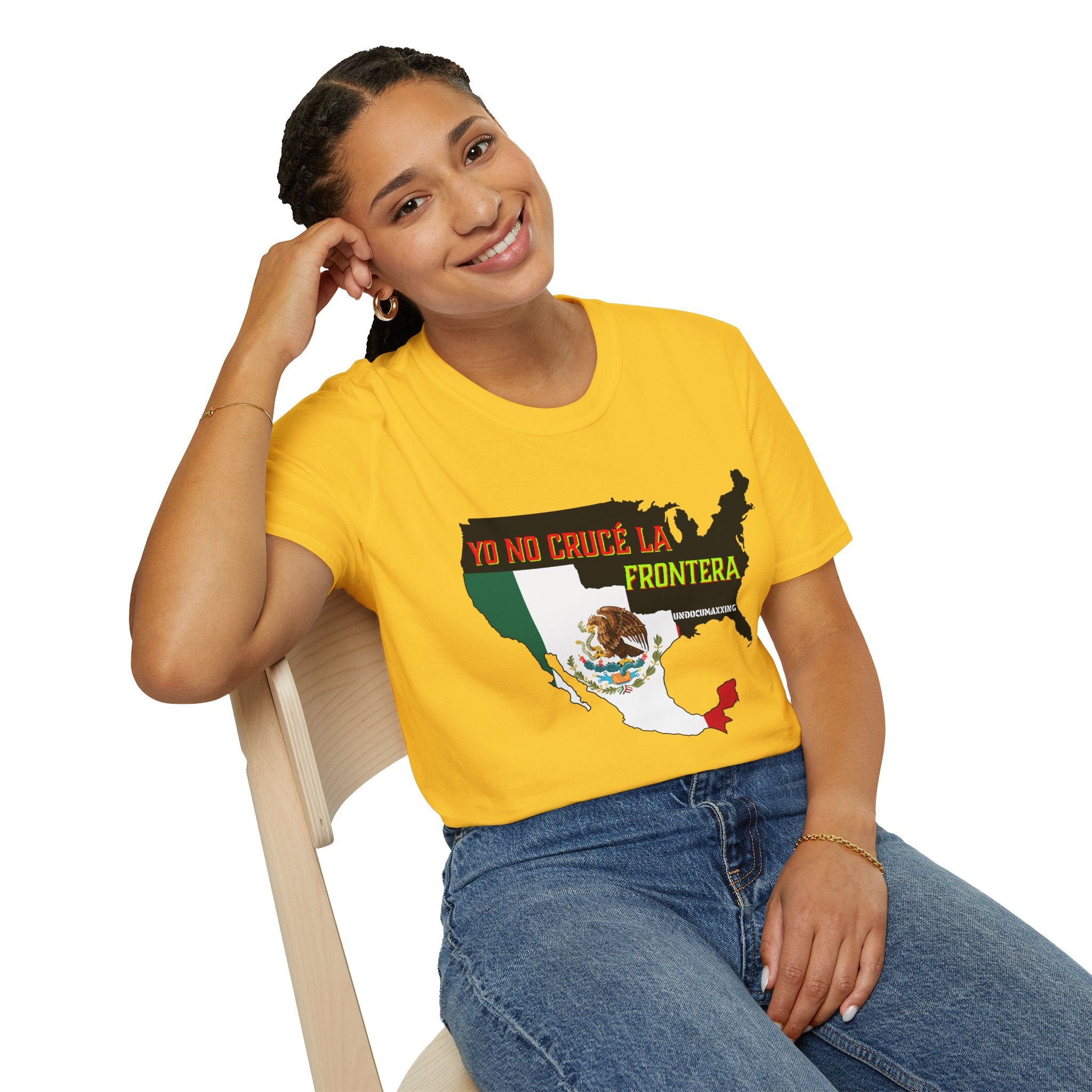 Daisy t-shirt A woman wearing a graphic t-shirt and blue jeans, sitting on a light-colored wooden chair against a solid white background. The t-shirt features a graphic of a map of the United States filled with the Mexican flag colors and a central eagle emblem, with the text 'YO NO CRUCE LA FRONTERA'.